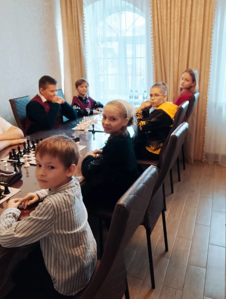 Дети сидят за длинным столом с шахматными досками