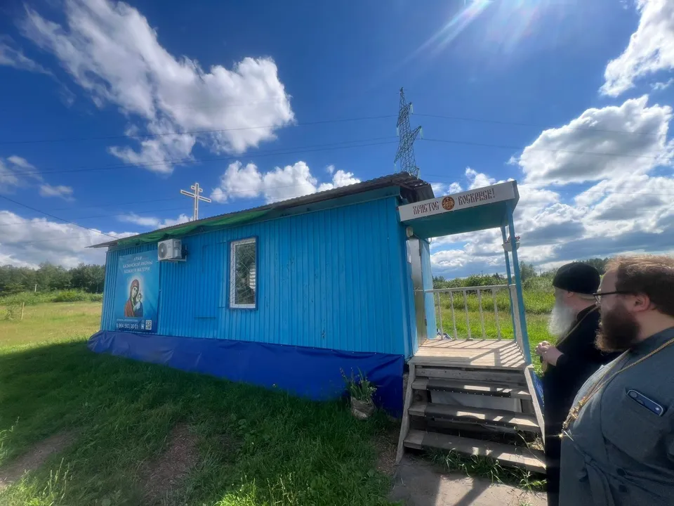 Синий вагончик-молитвенный дом с крестом на крыше, баннером иконы и крыльцом; у входа стоят два священнослужителя